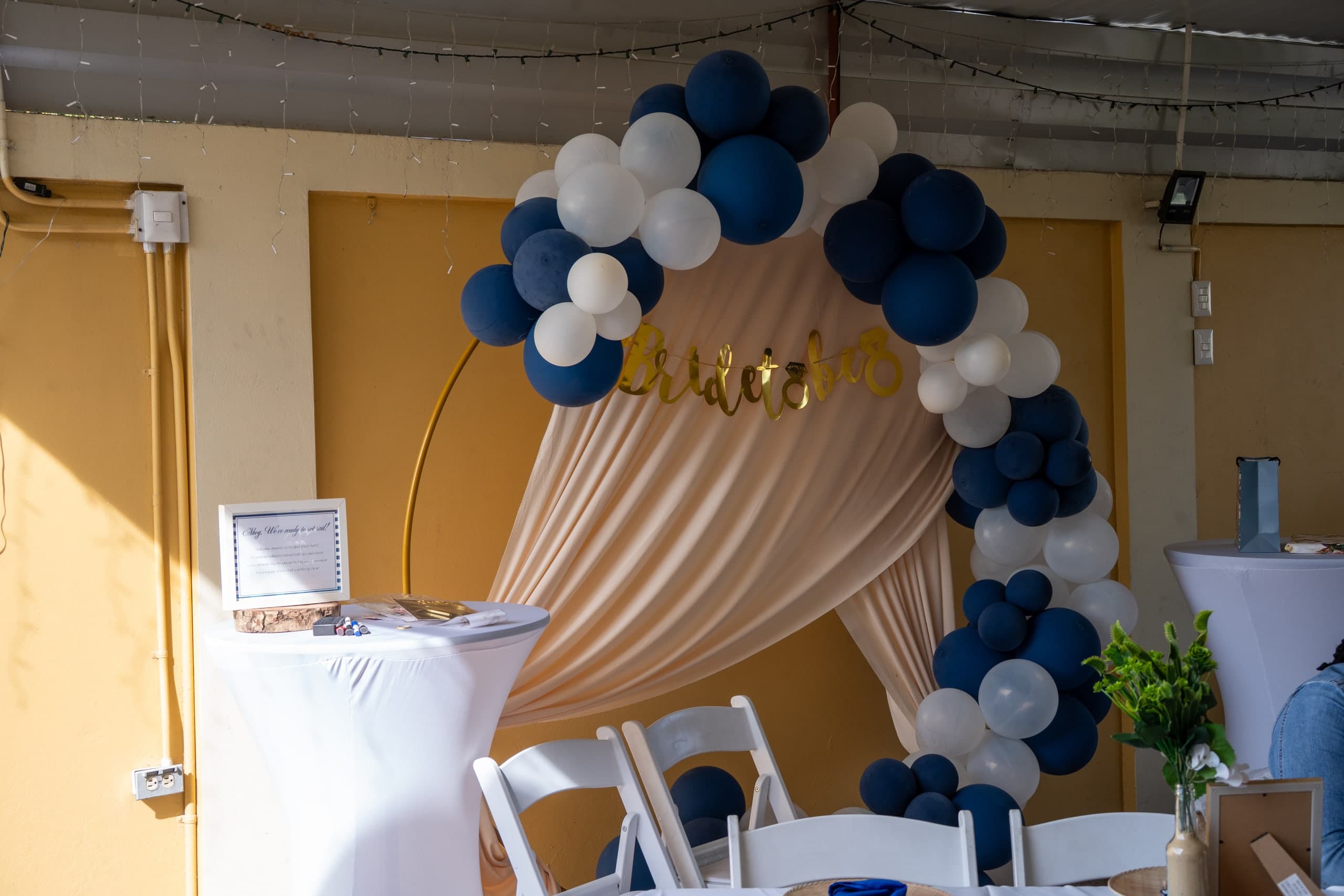 A navy and white balloon arch around a gold hoop, framing blush draping and a gold script sign, with white chairs and cocktail tables in front.
