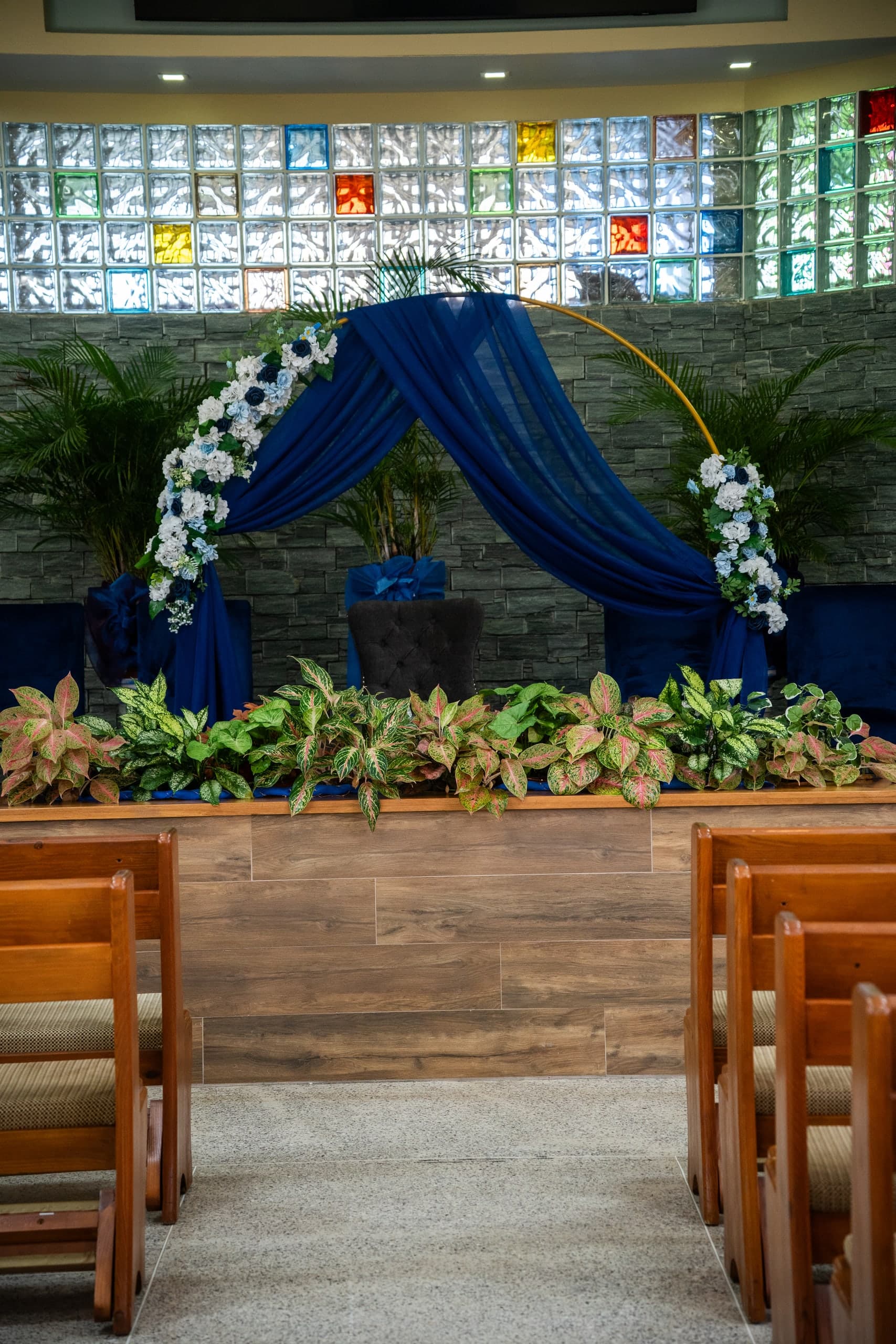 A gold circular arch draped in royal blue fabric with white and blue florals, set behind a planter of tropical foliage.