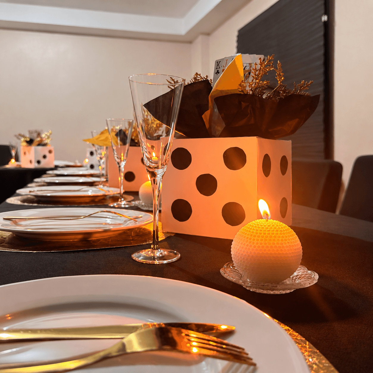 A black table set with gold chargers, white plates, gold cutlery and champagne flutes, with a lit candle and white dice-style centrepieces.