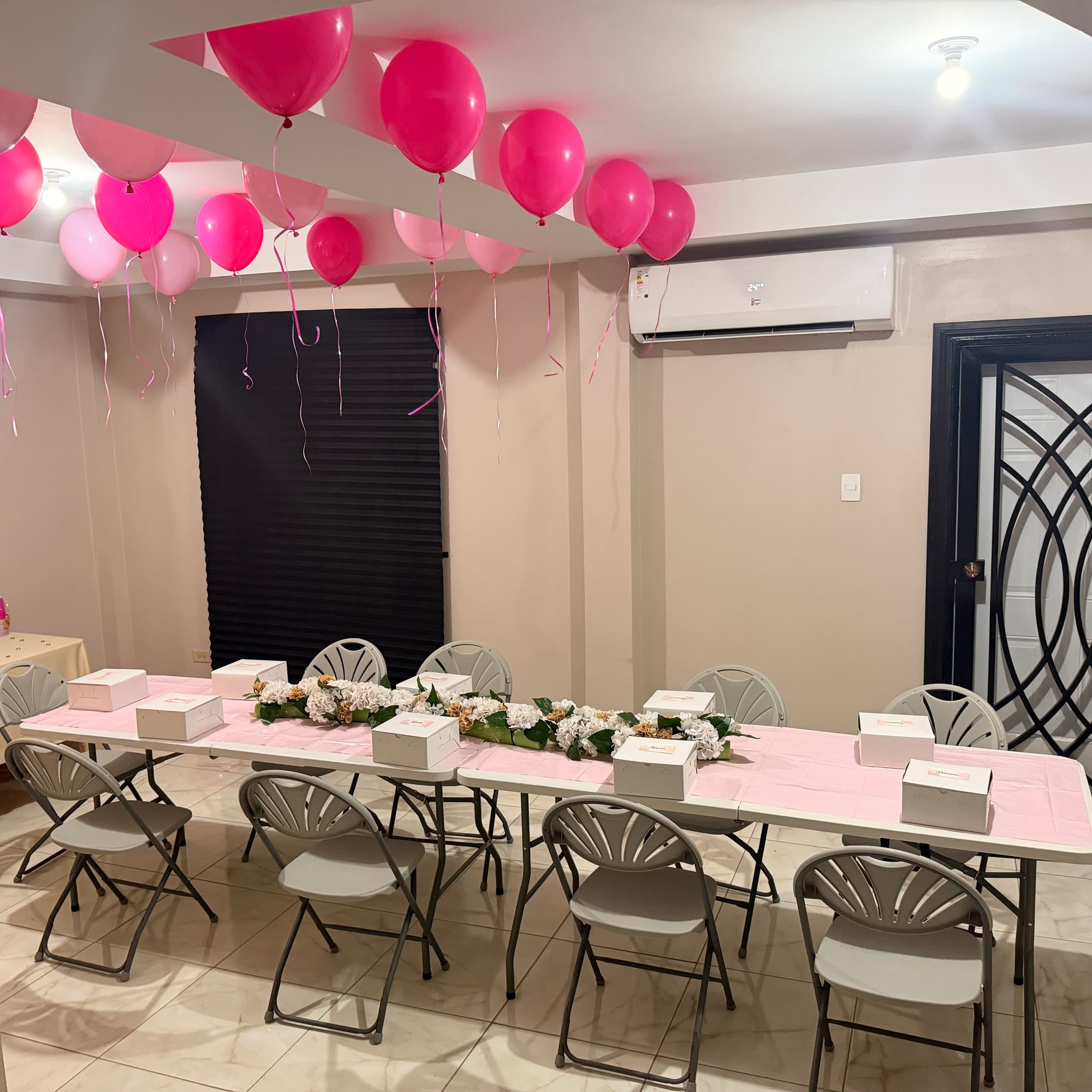 A long table dressed in pink with white boxes at each place and a runner of white hydrangeas, beneath pink balloons floating at the ceiling.