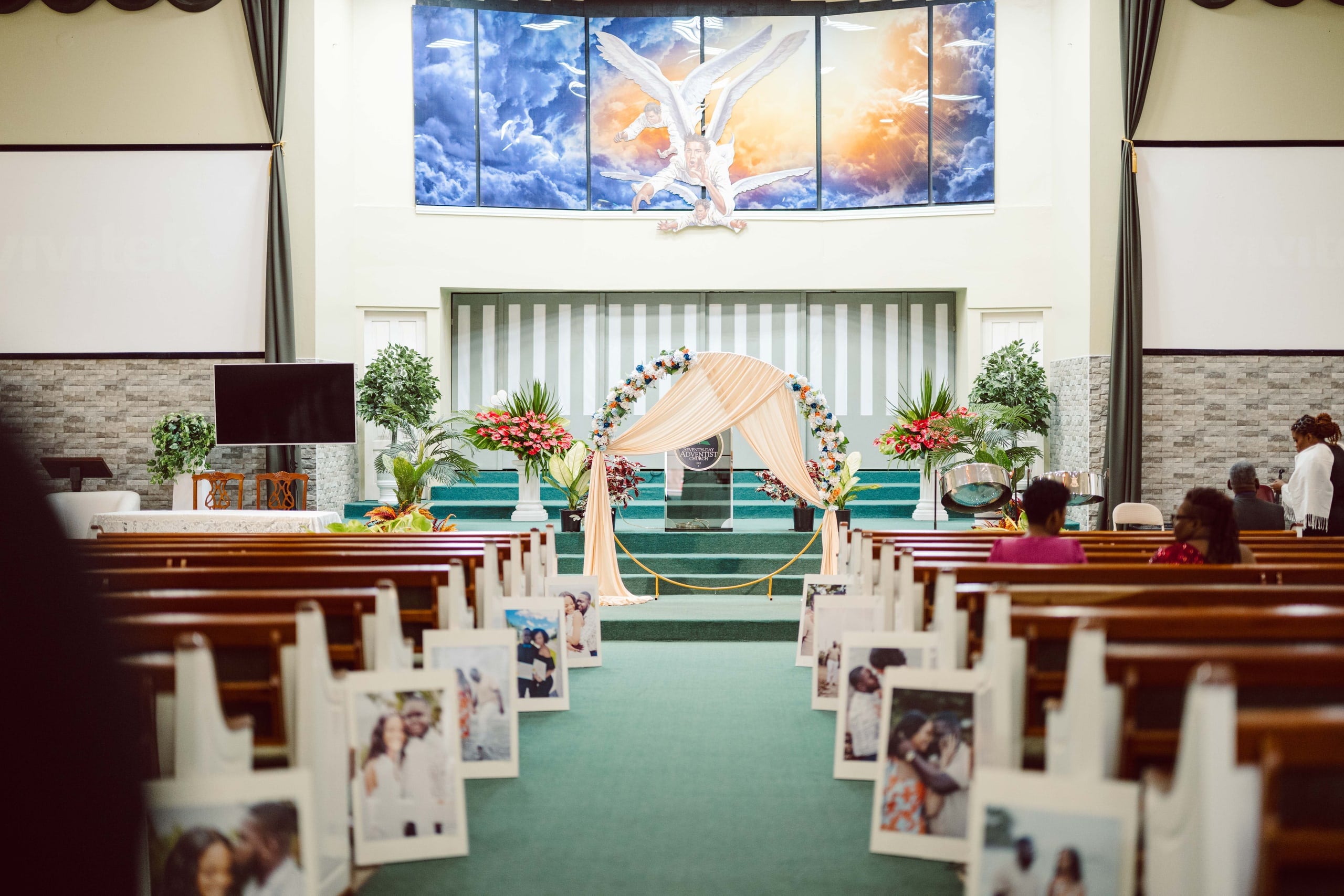 A church sanctuary with a champagne draped arch topped in florals, framed by a stained-glass angel backdrop, with guest photographs lining the pews.